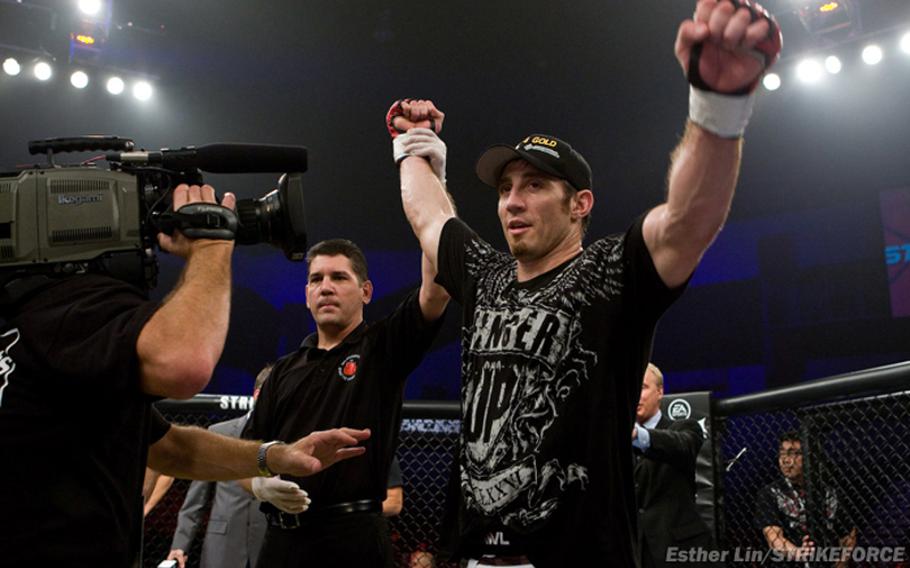 A man in a black T-shirt and hat raises an arm in celebration as a referee raises the other inside a mixed martial arts ring.